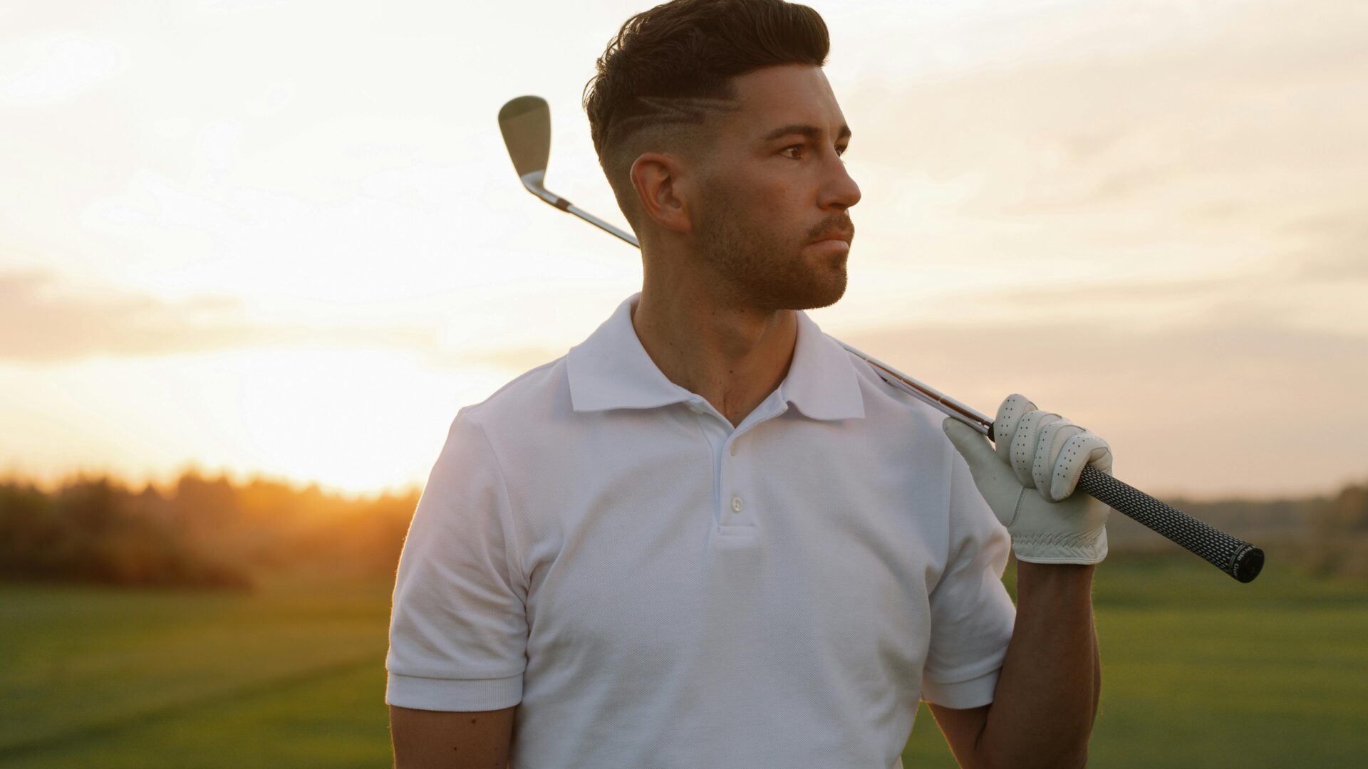 Joueur de golf sur un terrain de golf, avec l'air déterminé. Il est en contrejour au coucher du soleil, porte un polo et tient un club de golf