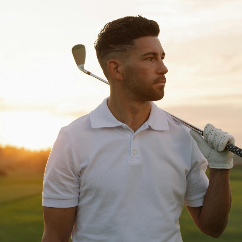 Joueur de golf sur un terrain de golf, avec l'air déterminé. Il est en contrejour au coucher du soleil, porte un polo et tient un club de golf